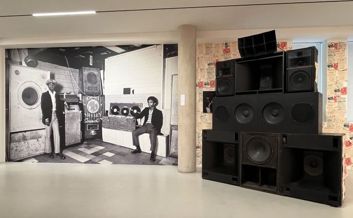 A l'exposition "Colored Black" de Dennis Morris à la Galerie du Jour, une photo en mode "wall paper" de Count Shelly, qui importa la culture jamaïcaine du sound system à Londres, ici en 1973, à côté d'un "mur d'enceintes" de sound system. (LAURE NARLIAN / FRANCEINFO)